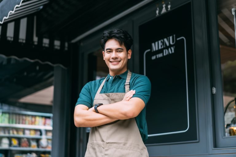 Young adult Asian business man crossing her arms in front of cafe restaurant while smiling. Happy and joyful barista of Coffee shop having arms crossed. High quality photo